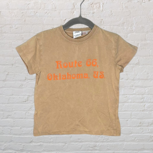 Tan Zara T-shirt with 'Route 66, Oklahoma, US' printed in orange, hung against a white brick wall.