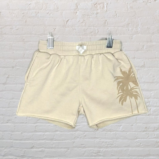 Beige sweat shorts featuring a palm tree design, displayed against a brick wall.