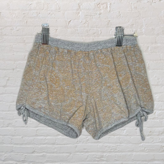 Heathered gray and tan shorts with drawstring details hanging on a clip against a white brick wall.