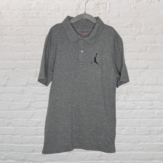 Grey Jordan polo shirt with embroidered Jumpman logo, hanging on a hanger against a white brick background.