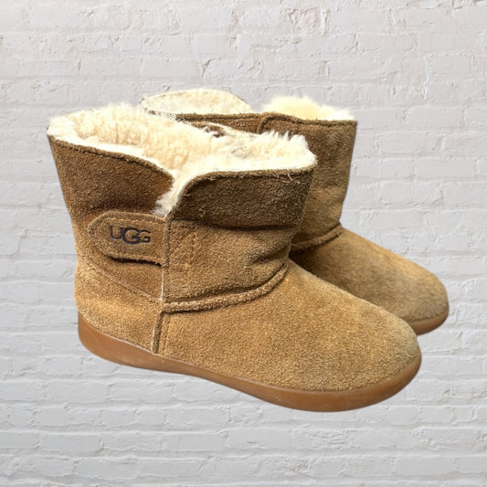 Chestnut UGG boots with shearling lining, featuring a suede upper and rubber sole, against a white brick background.