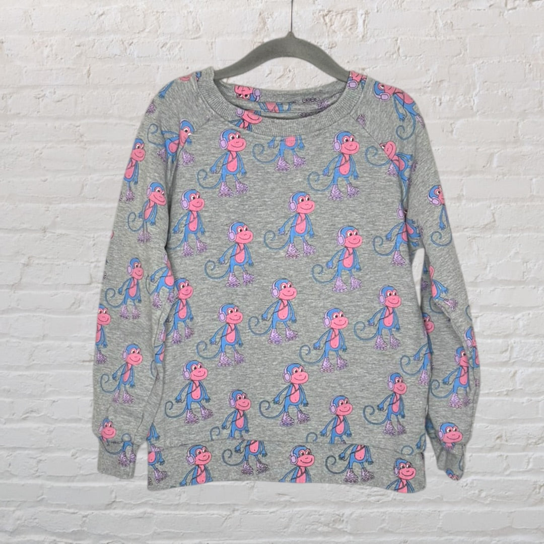 Grey sweatshirt with a playful pink and blue monkey pattern, hanging on a hanger against a white brick wall.