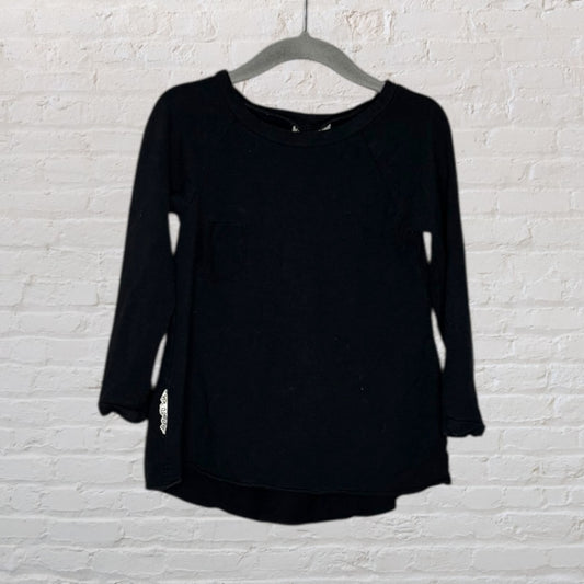 Black long sleeve tunic with a simple design, hanging on a grey hanger against a white brick wall.