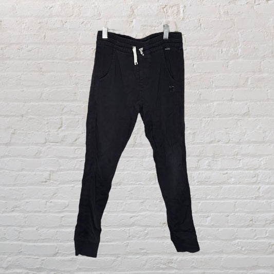 Black jogger pants with drawstring waist and side pockets, hanging against a white brick wall.