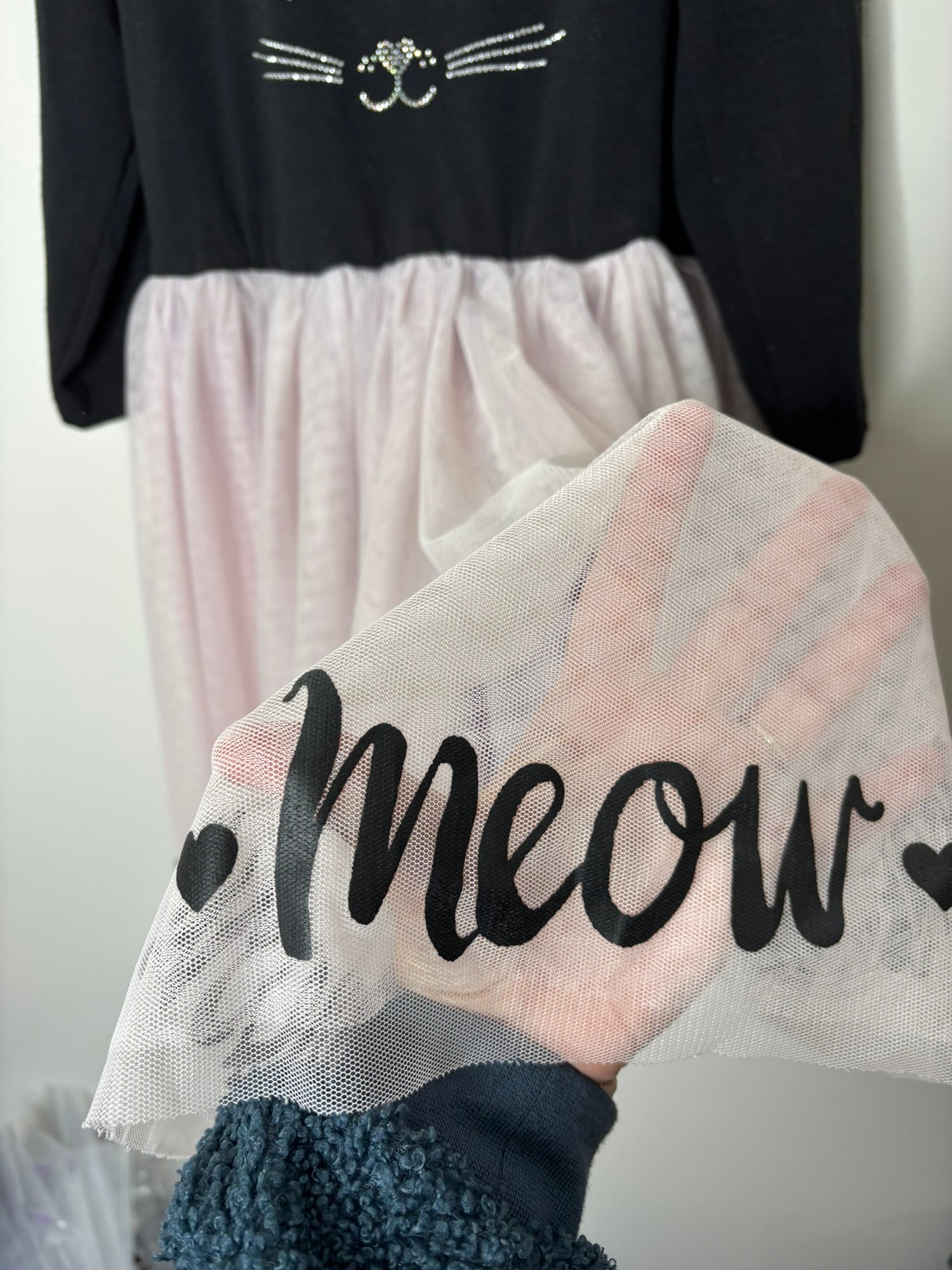 Black long sleeve dress with cat face design and pink tulle skirt, hanging on a hanger against a white brick wall.