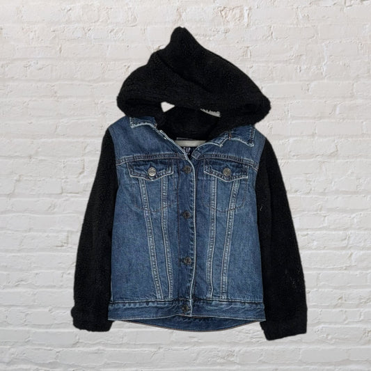 Denim jacket with black sherpa fleece sleeves and hood, hanging against a white brick wall.