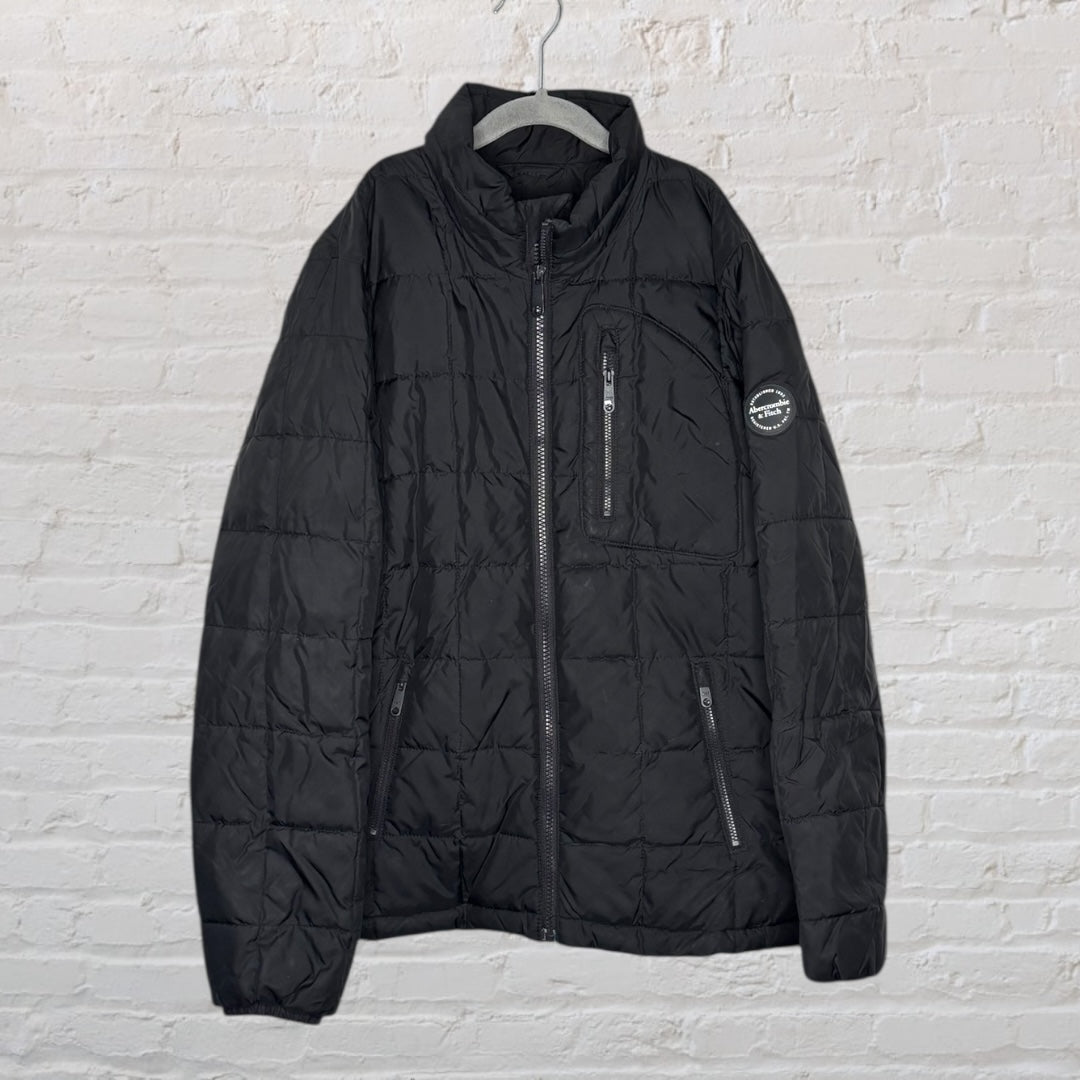 Black quilted puffer jacket with zipper pockets and a round patch on the sleeve, hanging against a white brick wall.