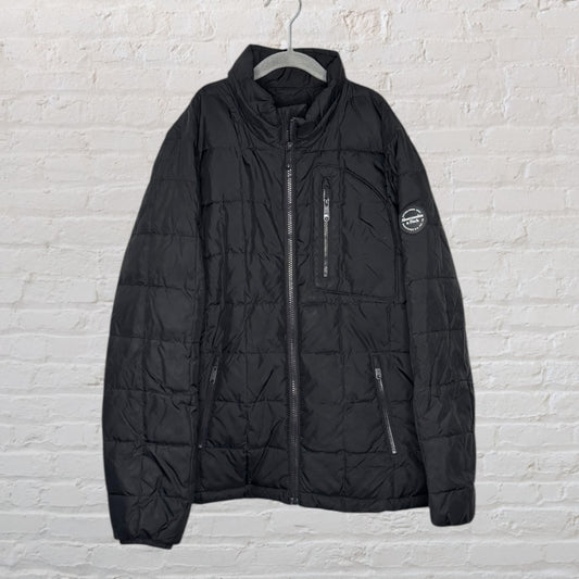 Black quilted puffer jacket with zipper pockets and a round patch on the sleeve, hanging against a white brick wall.
