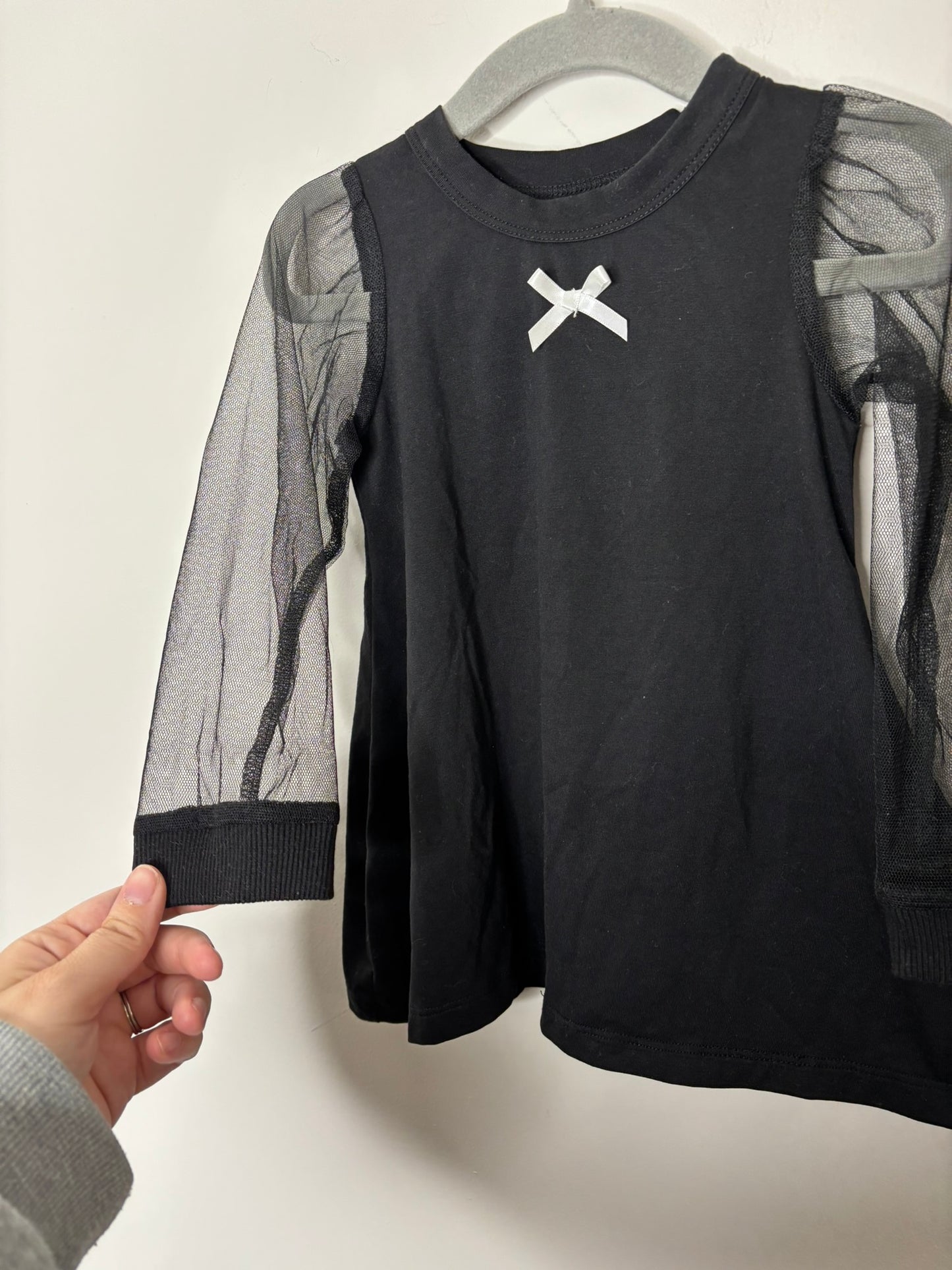 Black top with mesh sleeves and a white bow detail at the neckline, hanging on a grey hanger.