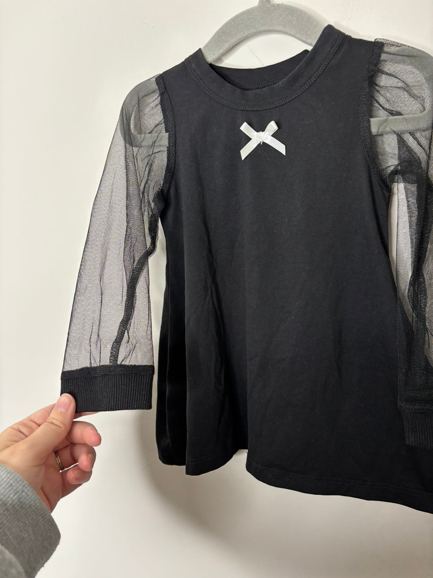 Black top with mesh sleeves and a white bow detail at the neckline, hanging on a grey hanger.