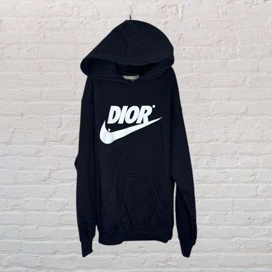 Black hoodie with a large white Dior logo and swoosh design, displayed against a brick wall.