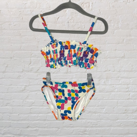 A colorful two-piece swimsuit with smocked top and vibrant geometric print bottom, hanging on a hanger.