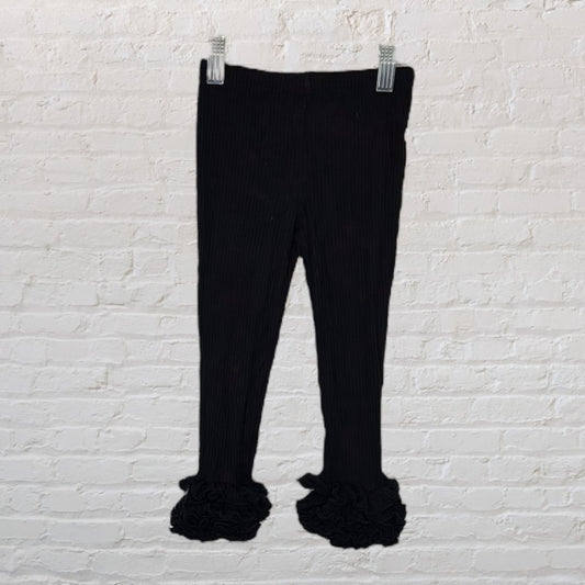 Black ribbed leggings with ruffled hem, hanging against a white brick wall background.