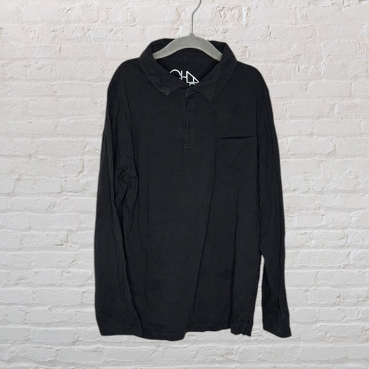 Black long sleeve polo shirt with pocket, hanging against a white brick wall.