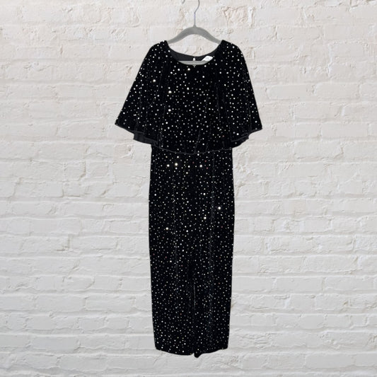 Black velvet jumpsuit with silver embellishments and a cape detail hanging on a hanger.