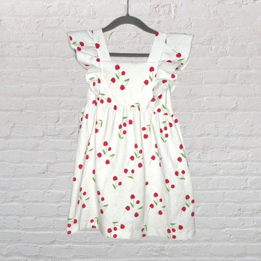 White dress with red cherry print and ruffled sleeves hanging on a black hanger against a textured wall.