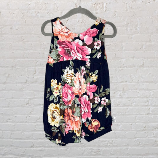 A sleeveless navy romper with vibrant floral print in pink, yellow, and green, hanging on a gray hanger against a brick wall.