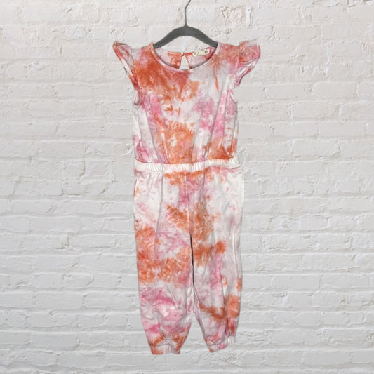 A tie-dye jumpsuit with pink and orange patterns, featuring ruffle sleeves, hanging on a hanger against a white brick background.