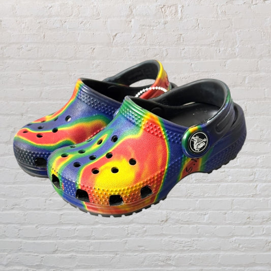 Pair of multicolor tie-dye Crocs clogs against a brick wall background.