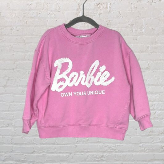 Pink Barbie sweater with 'Barbie Own Your Unique' text in white, displayed on a hanger against a white brick wall.