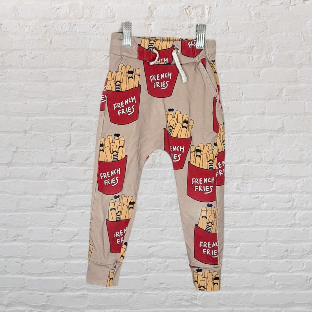 Beige jogger pants with a playful French fries graphic print, featuring a drawstring waist, hanging on clips against a white brick wall.
