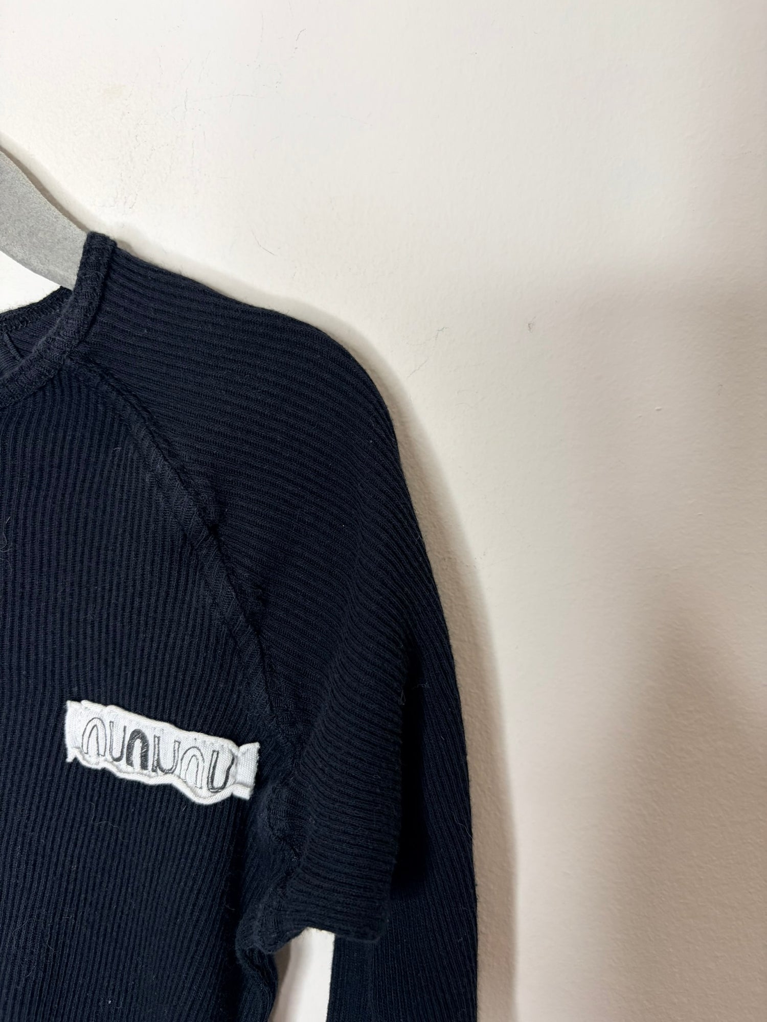 Black long sleeve shirt with Nununu branding on the chest and hem, hung on a grey hanger against a white brick wall.