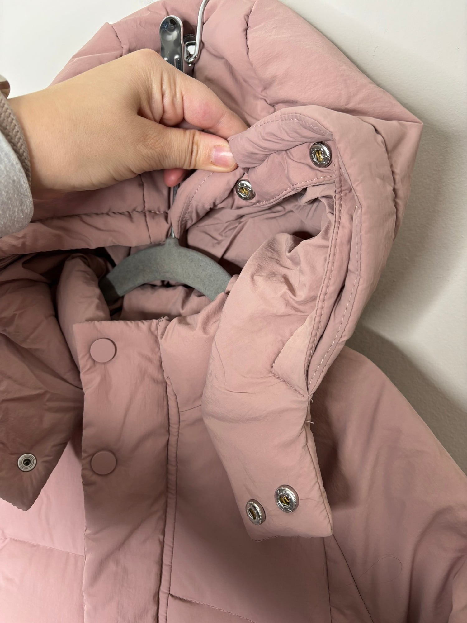 Pink hooded puffer jacket with front snap buttons, displayed on a hanger against a brick wall.