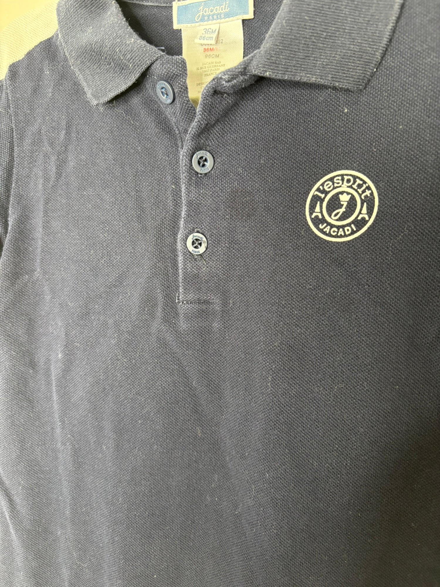 Black polo-style romper with white sleeves and a small embroidered logo, hanging on a hanger against a brick wall.