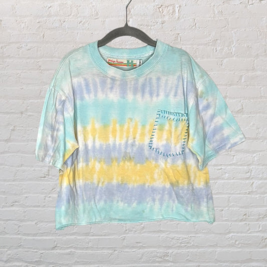 Cropped tie-dye t-shirt in blue, yellow, and white with a pocket, hanging on a hanger against a brick wall.