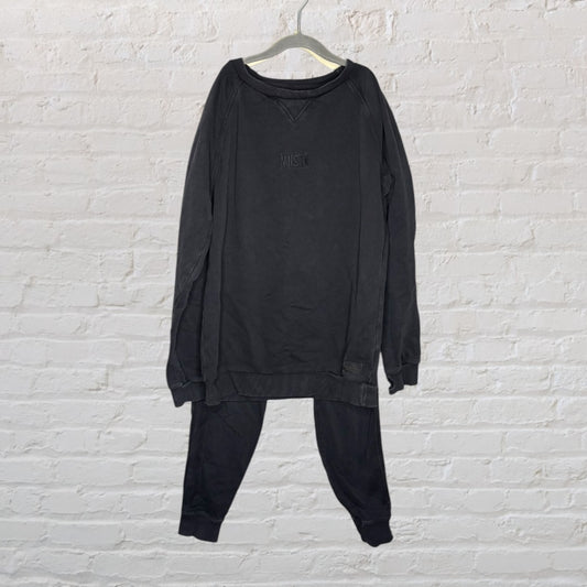 Black lounge set including a sweatshirt and pants on a hanger against a white brick wall.