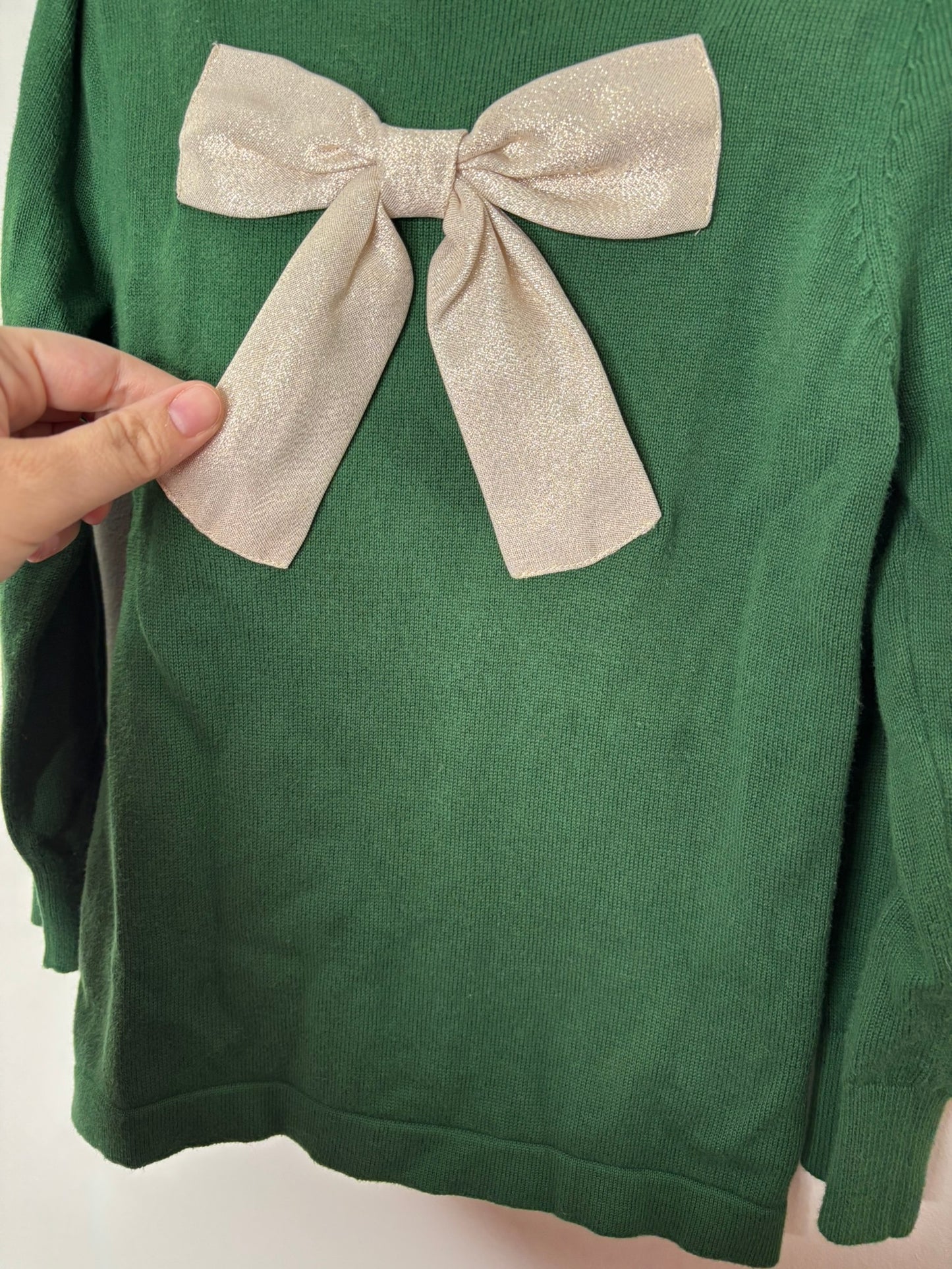 Green sweater with a large decorative beige bow on the front, hanging on a hanger against a white brick background.