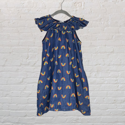 Denim dress with colorful rainbow prints and ruffled neckline, hanging on a hanger against a white brick wall.