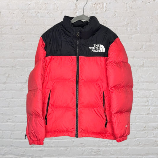 Red and black The North Face puffer jacket with logo detail on the chest, hanging on a white brick wall.