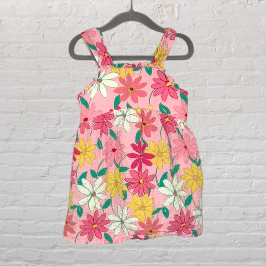 A pink sleeveless sundress with a multicolored floral pattern, hanging against a white brick wall.