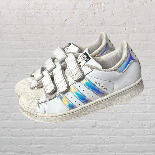 Pair of white Adidas Superstar sneakers with Velcro straps and iridescent stripes against a brick wall.