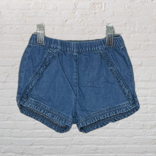 Blue denim shorts with ruffle detailing and elastic waist displayed on a hanger against a brick wall.