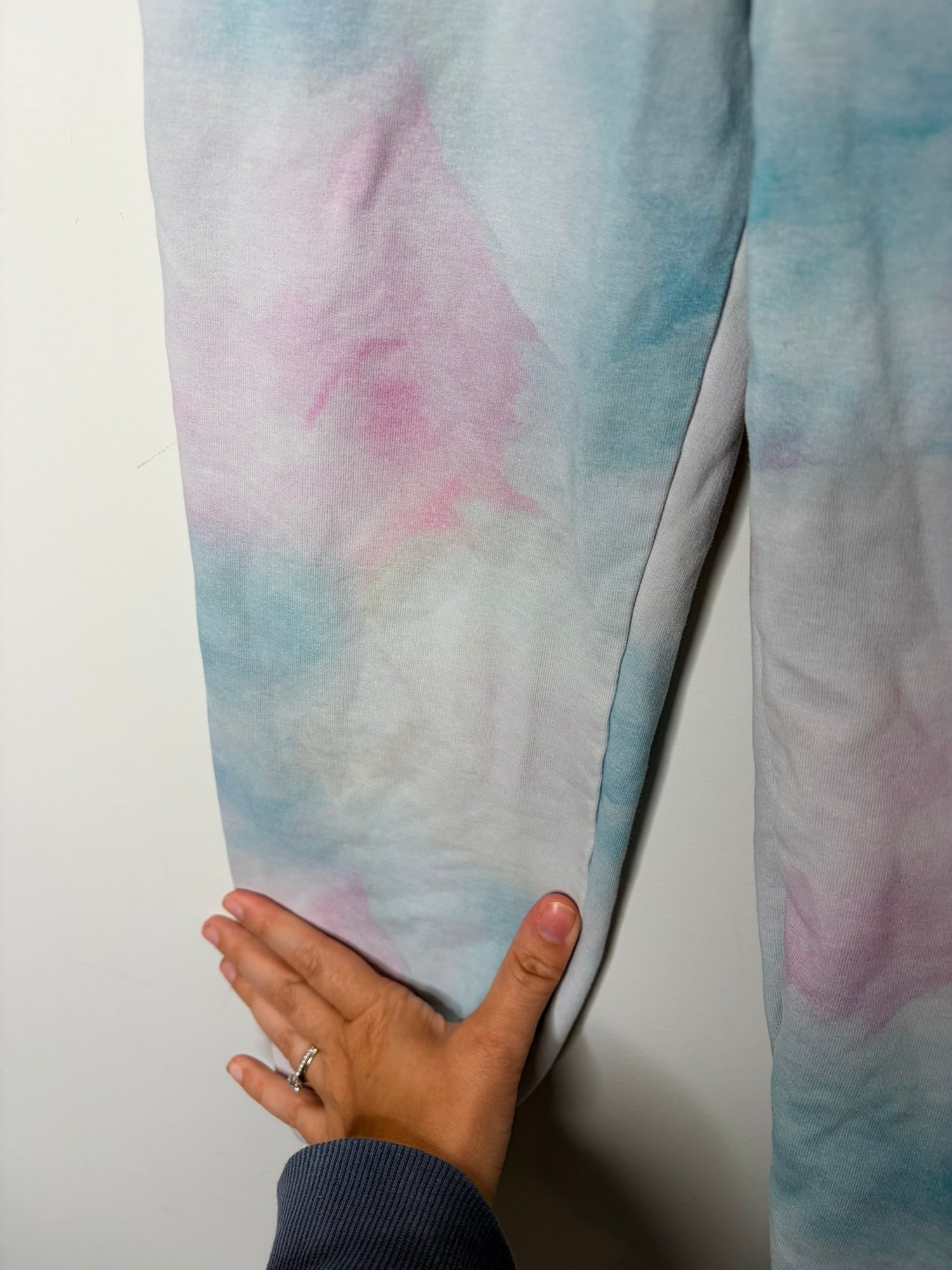 Unisex pastel tie-dye jogger pants in shades of pink, blue, and white, hanging on white clips against a white brick wall.