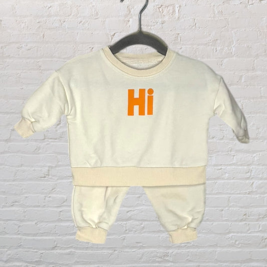 Cream baby outfit set with orange 'Hi' print, featuring a top and matching pants on a hanger.