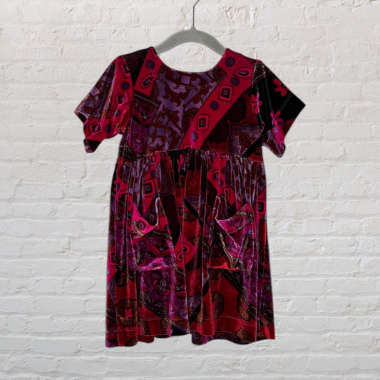 A red and purple velvet dress with a paisley pattern and short sleeves, hanging on a hanger against a brick wall.