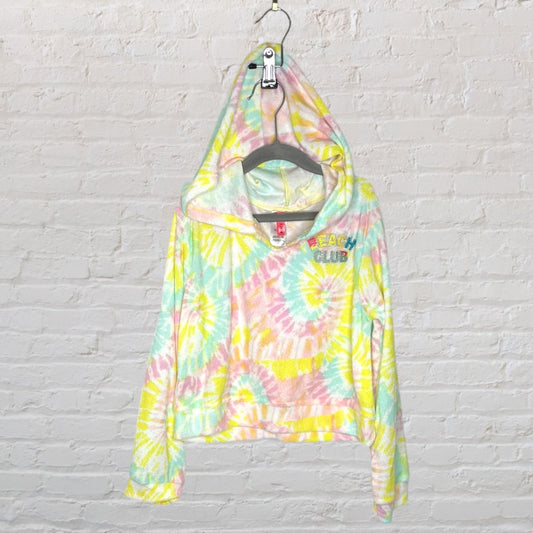 Pastel tie-dye hoodie with 'Beach Club' embroidery, hanging on a hanger against a white brick wall.