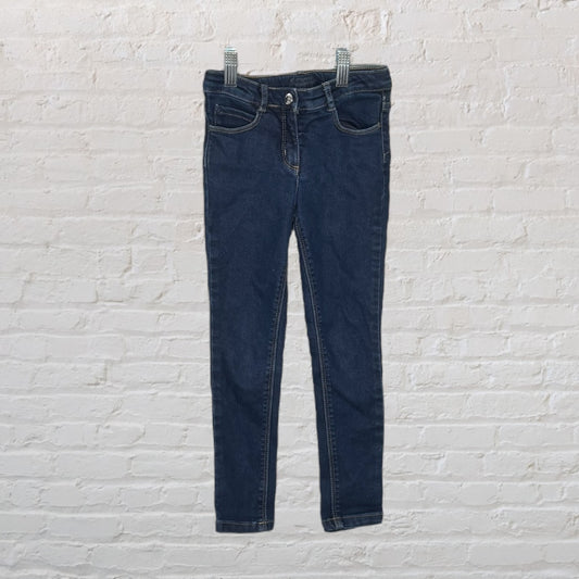 Children's dark blue skinny jeans with classic pocket design displayed against a white brick wall.