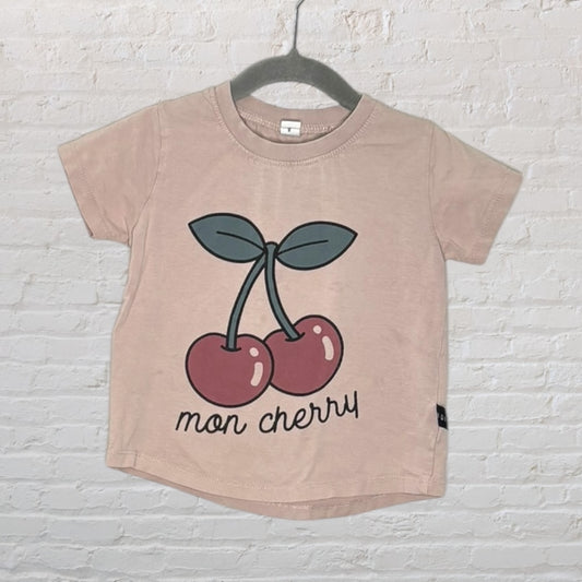 Light pink T-shirt with a graphic of two cherries and 'mon cherry' text, displayed on a hanger against a brick wall.