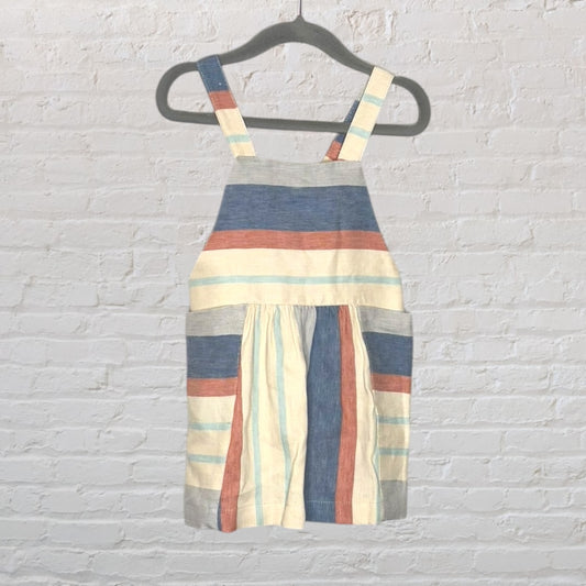 A multi-colored striped pinafore dress with shoulder straps and a gathered waist, hanging on a hanger against a brick wall.