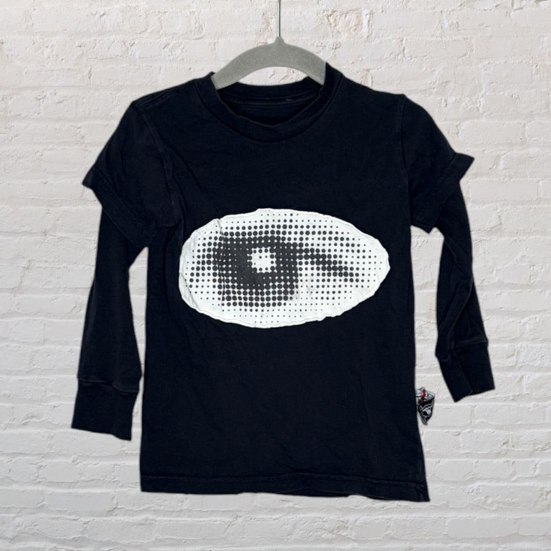 Black long sleeve t-shirt with a large white graphic eye design on the front, hanging against a brick wall backdrop.