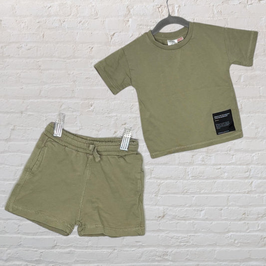 Kids olive green T-shirt with patch and matching shorts, displayed against a white brick wall.