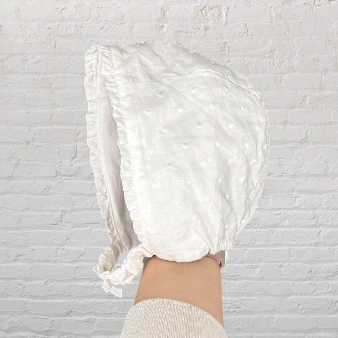 White vintage-style baby bonnet with delicate embroidery, held in front of a brick wall background.