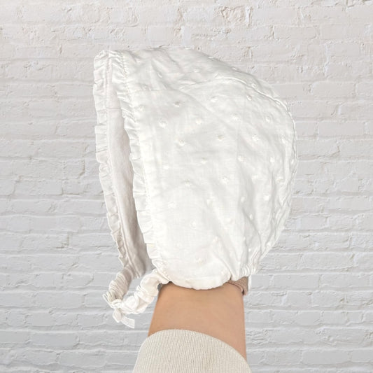 White vintage-style baby bonnet with delicate embroidery, held in front of a brick wall background.