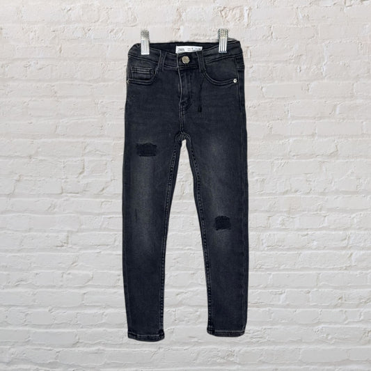 Black distressed skinny jeans for kids by Zara, displayed on hangers against a white brick wall.