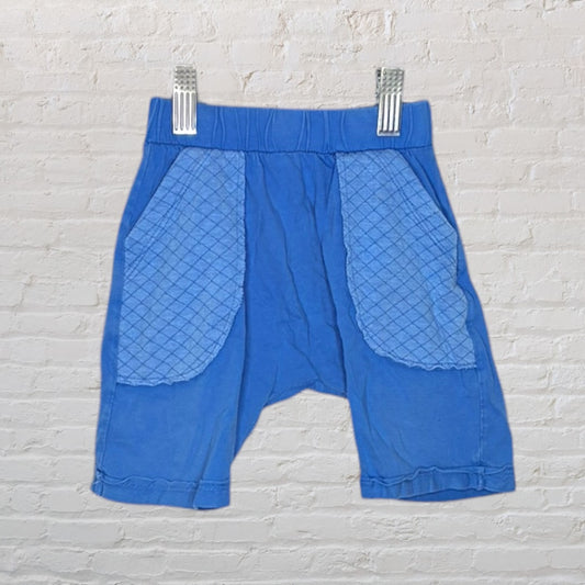 Blue kids shorts with quilted pockets, displayed on a hanger against a white brick background.