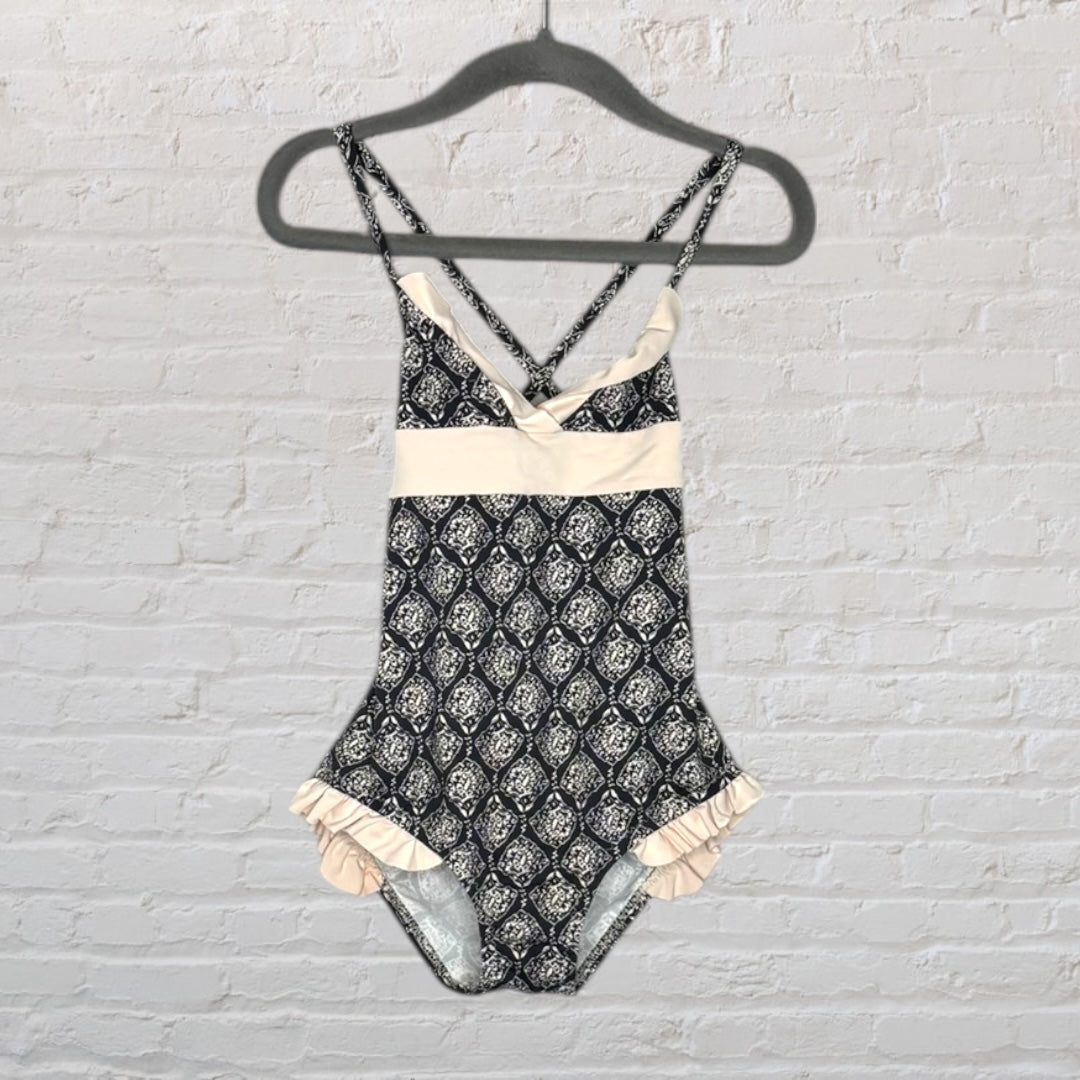 Women's one-piece swimsuit with a black and cream pattern, featuring ruffles and cross-back straps, hanging on a black hanger.